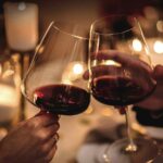 Due calici di vino rosso che si toccano per un brindisi romantico, con un'atmosfera intima e candele accese sullo sfondo.